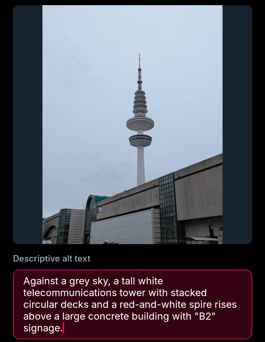 Against a grey sky, a tall white telecommunications tower with stacked circular decks and a red-and-white spire rises above a large concrete building with "B2" signage.